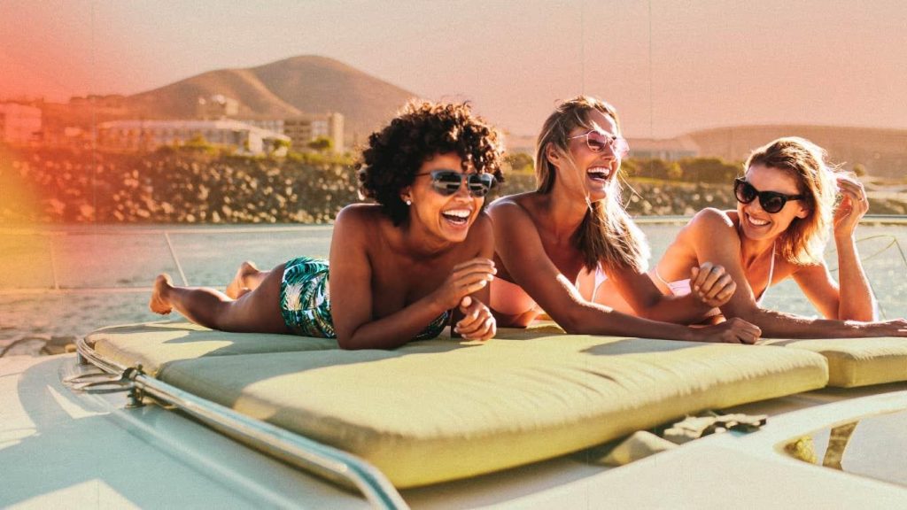 Three happy women relaxing and laughing on a yacht bow sun pad during golden hour